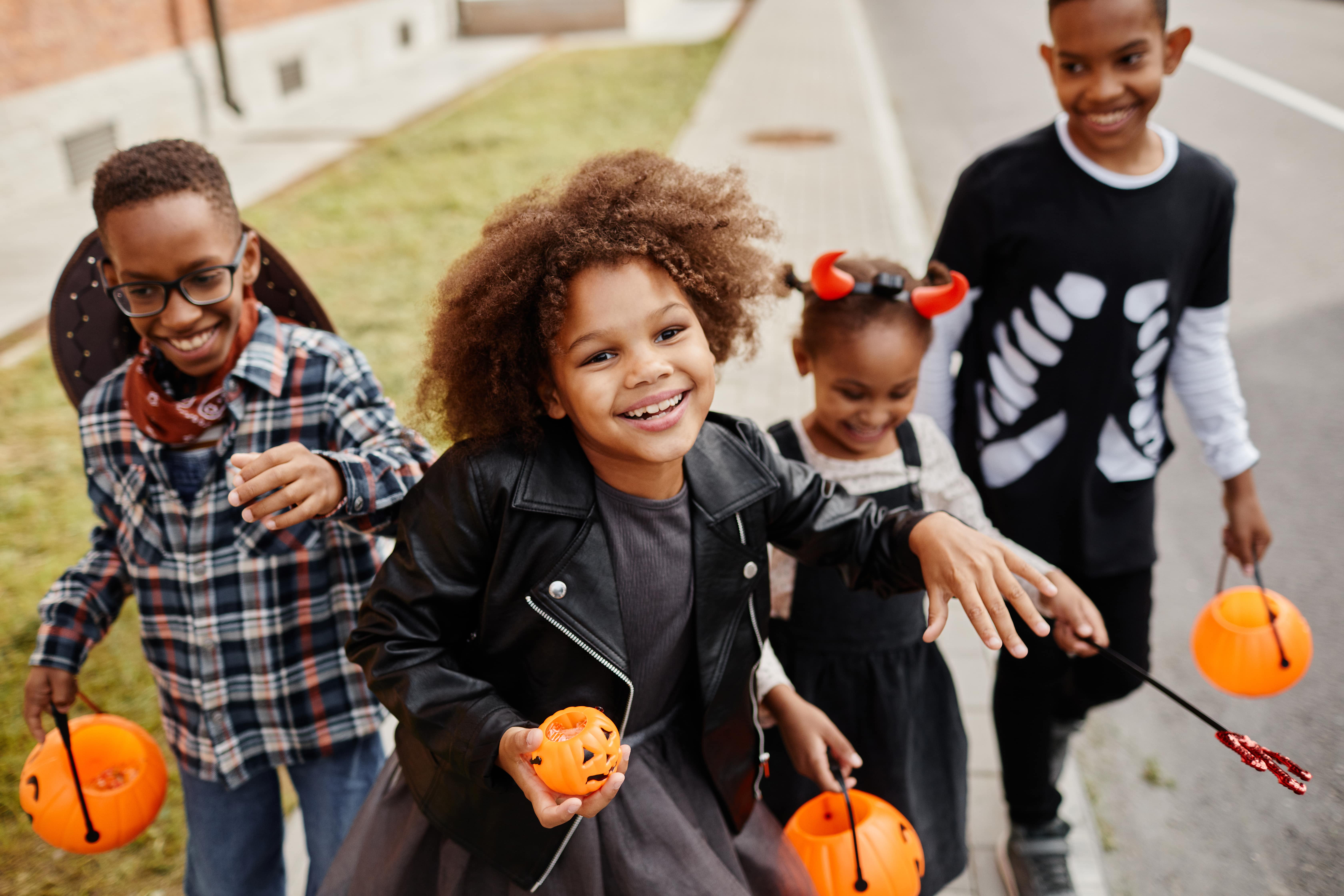 A group of young children walking on the sidewalk dressed in costumes holding pumpkin baskets for trick-or-treating. 