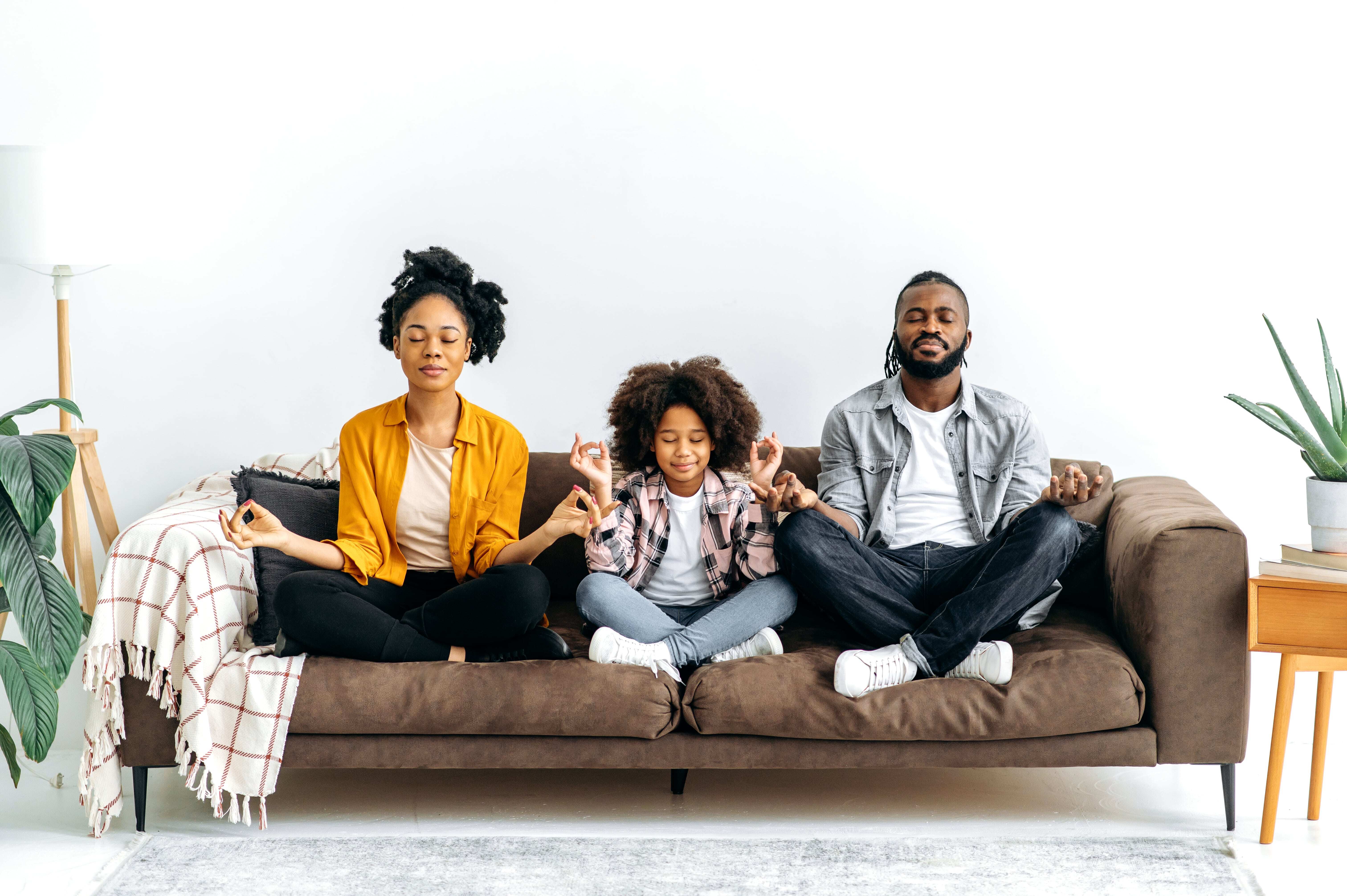 A young family of three sitting on the couch with their eyes closed, legs crossed, and hands on their knees practicing daily mindfulness activities for kids. 